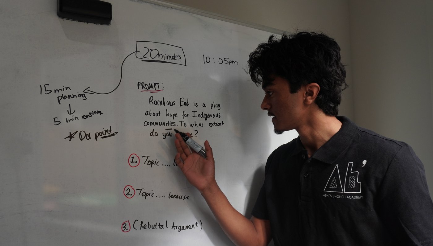 Abhi teaching a live VCE English lesson, breaking down an essay prompt on a whiteboard.